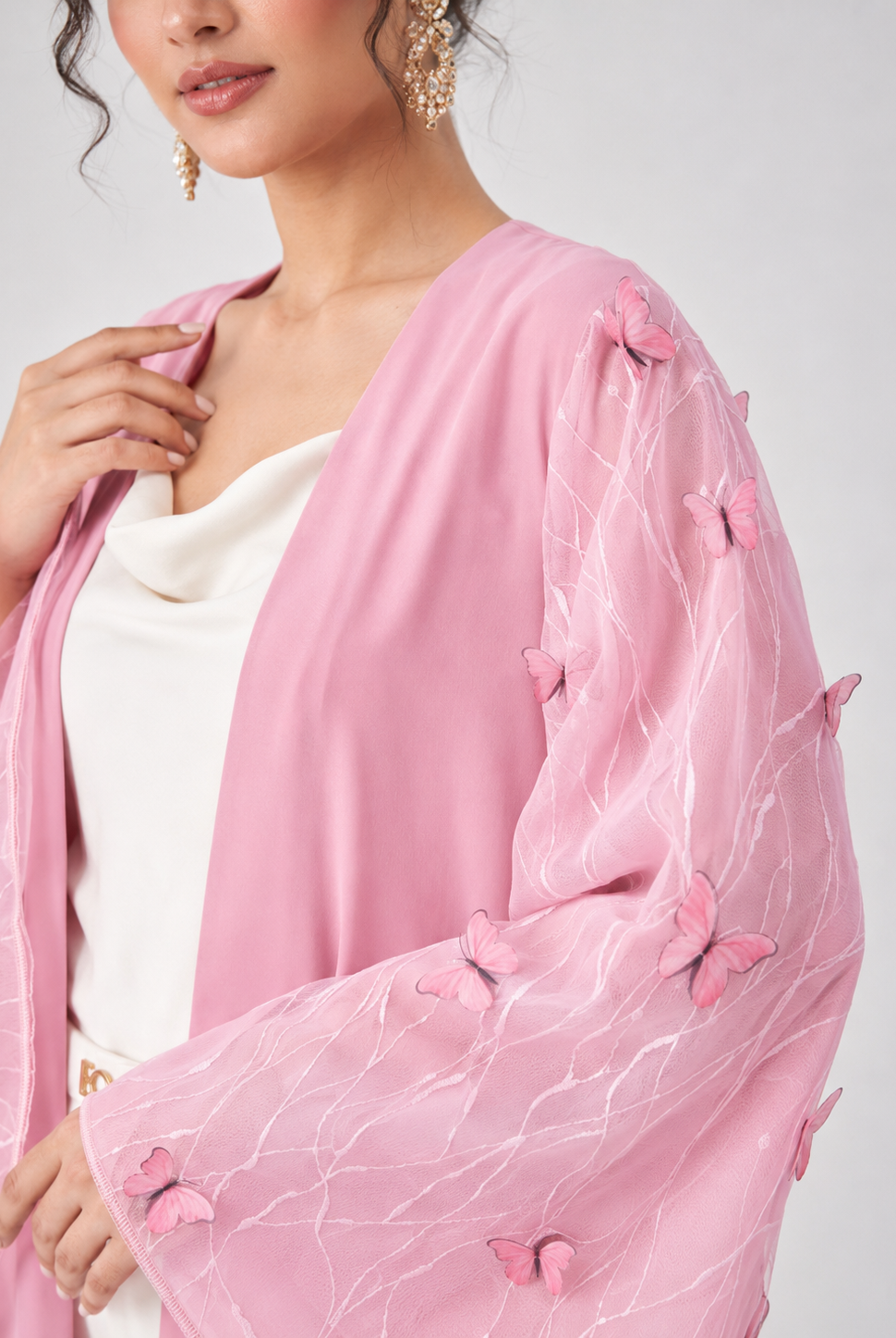 A close-up of a woman wearing a light pink open-front cardigan with sheer flutter sleeves decorated with 3D butterfly appliqués, layered over a white top against a light gray background.