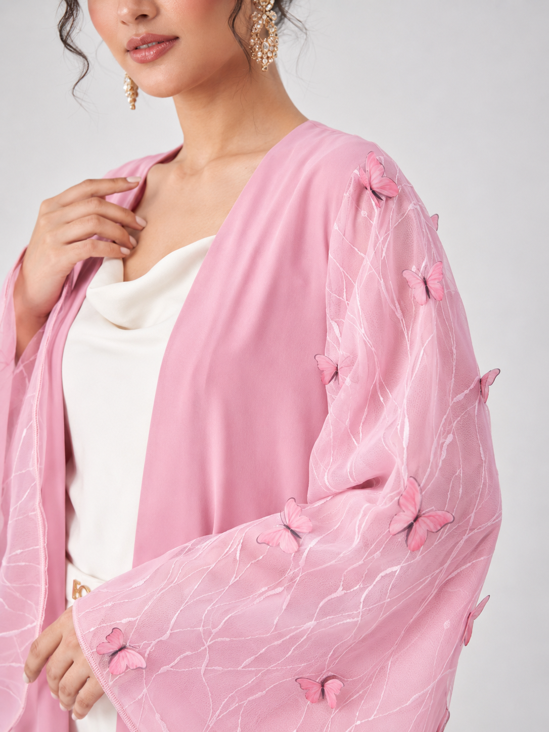 A close-up of a woman wearing a light pink open-front cardigan with sheer flutter sleeves decorated with 3D butterfly appliqués, layered over a white top against a light gray background.