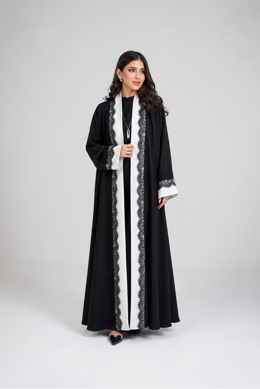 A woman modeling a long black abaya with white lace trim, worn over a black inner dress, against a neutral gray studio background.