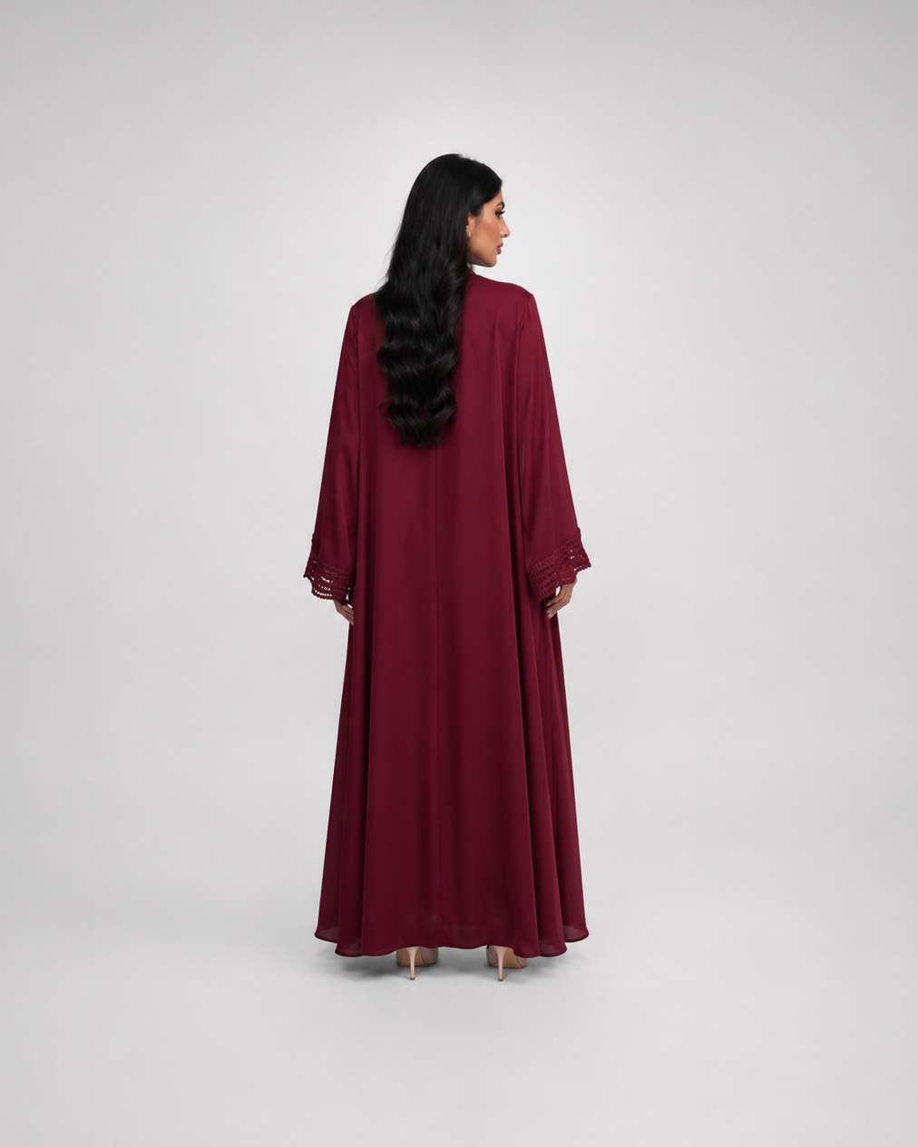 A woman with long black hair is shown from behind wearing a long, flowing maroon abaya with wide sleeves, standing against a light gray studio background.