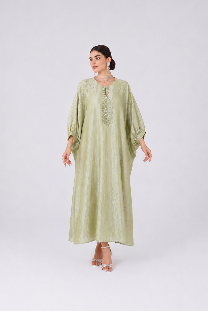 A woman modeling a long, flowing sage green dress with wide sleeves and embroidered detailing on the chest, standing against a light gray studio background.