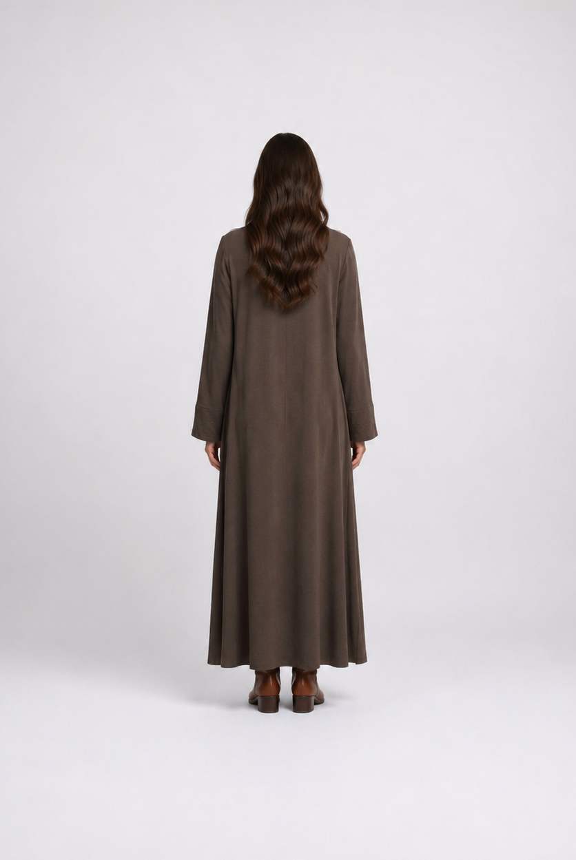 A woman viewed from behind wearing a long, loose-fitting brown maxi dress with wide sleeves, standing against a plain light gray studio background.