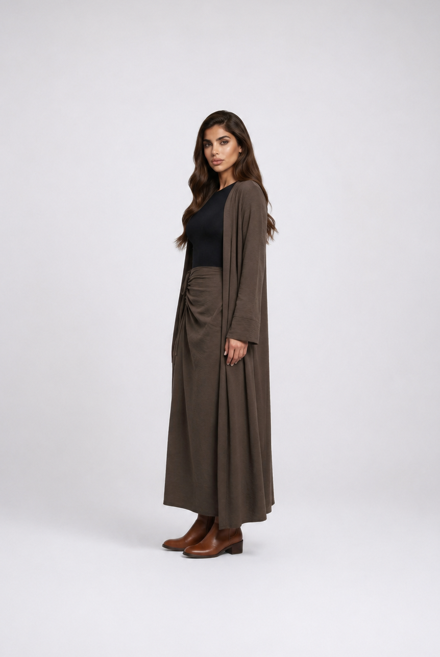 A woman modeling a long brown draped cardigan over a black top and matching brown pleated maxi skirt, standing against a light gray studio background.