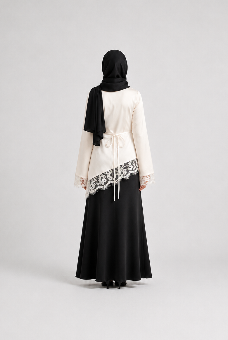 A woman viewed from behind wearing a black hijab, a cream-colored long-sleeve top with black lace trim, and a long black skirt, standing against a light gray studio background.