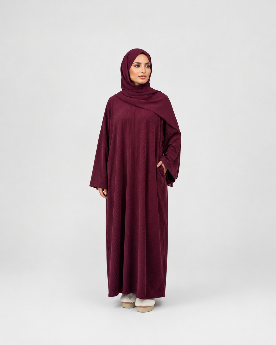 A woman modeling a long burgundy abaya with a matching hijab, standing against a light gray studio background.