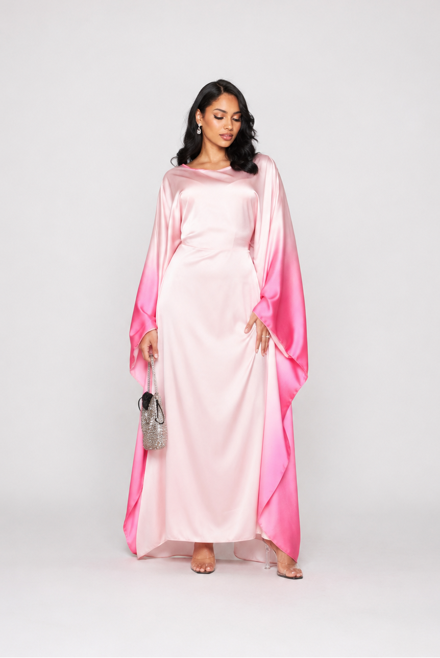 A woman modeling a long, flowing satin dress with a light pink body and vibrant pink draped sleeves, standing against a plain light gray studio background.