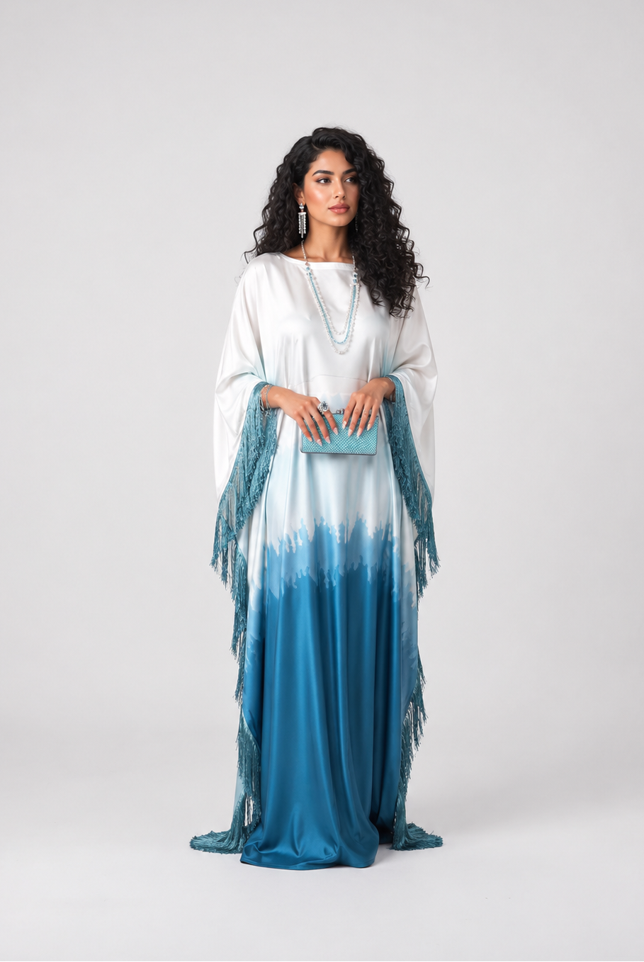 A woman modeling a long white and blue tie-dye maxi dress with fringe details on the sleeves, standing against a light gray studio background.