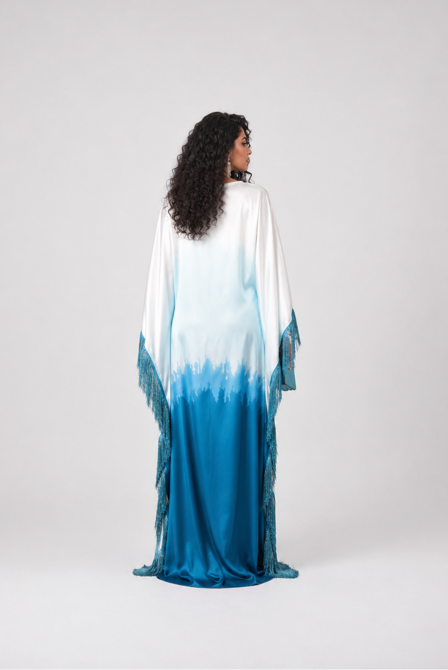 A woman stands with her back to the camera wearing a long flowing kaftan dress with a white-to-blue ombré gradient and fringe details, photographed against a light gray studio background.