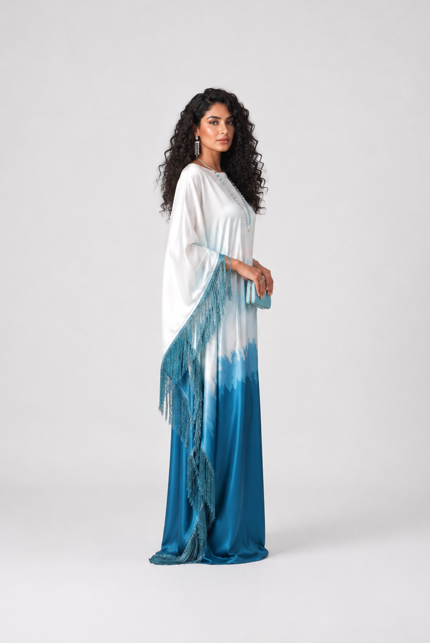 A woman modeling a long white and blue tie-dye maxi dress with fringe details, standing against a light gray studio background.