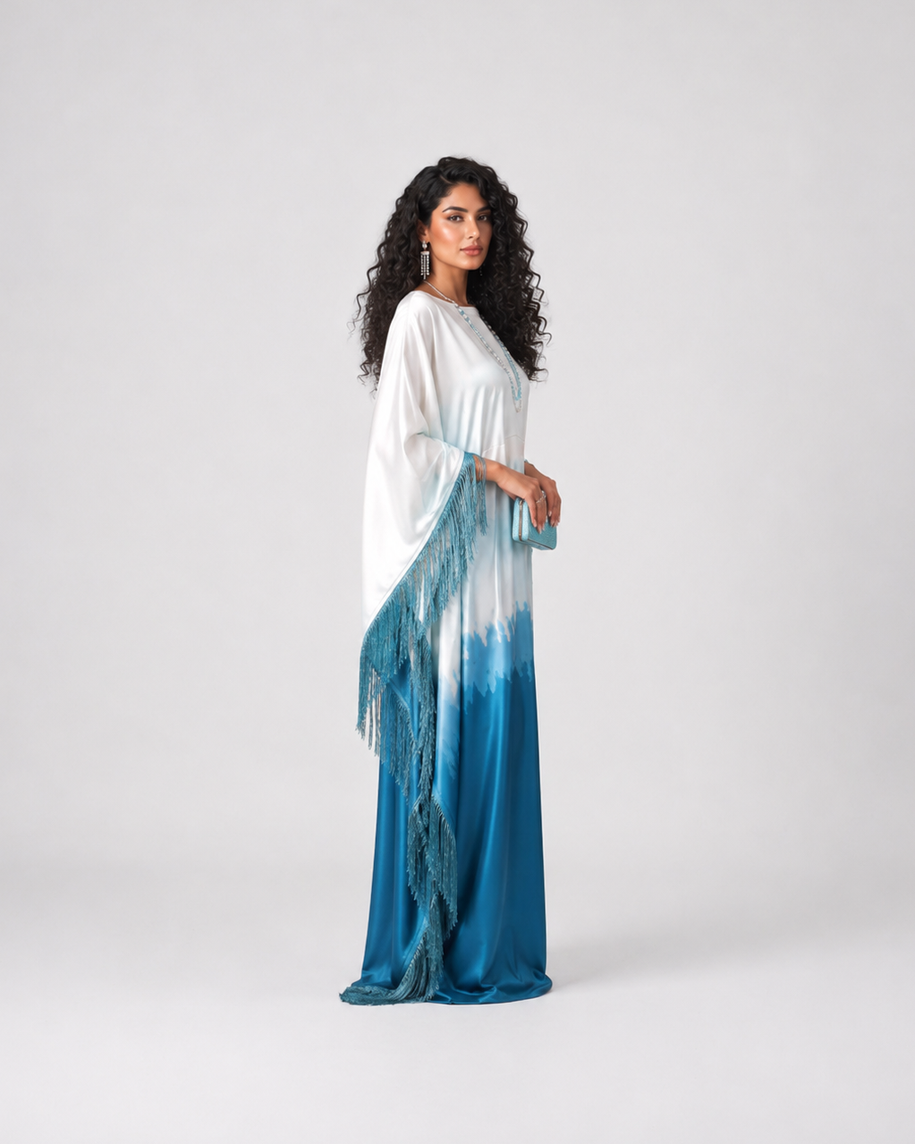 A woman modeling a long white and blue tie-dye maxi dress with fringe details, standing against a light gray studio background.