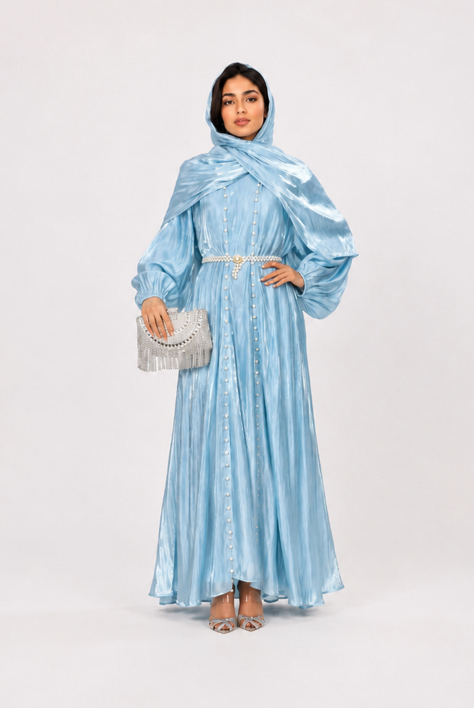 A woman modeling a light blue metallic maxi dress with a matching headscarf and holding a silver clutch, standing against a plain light gray studio background.