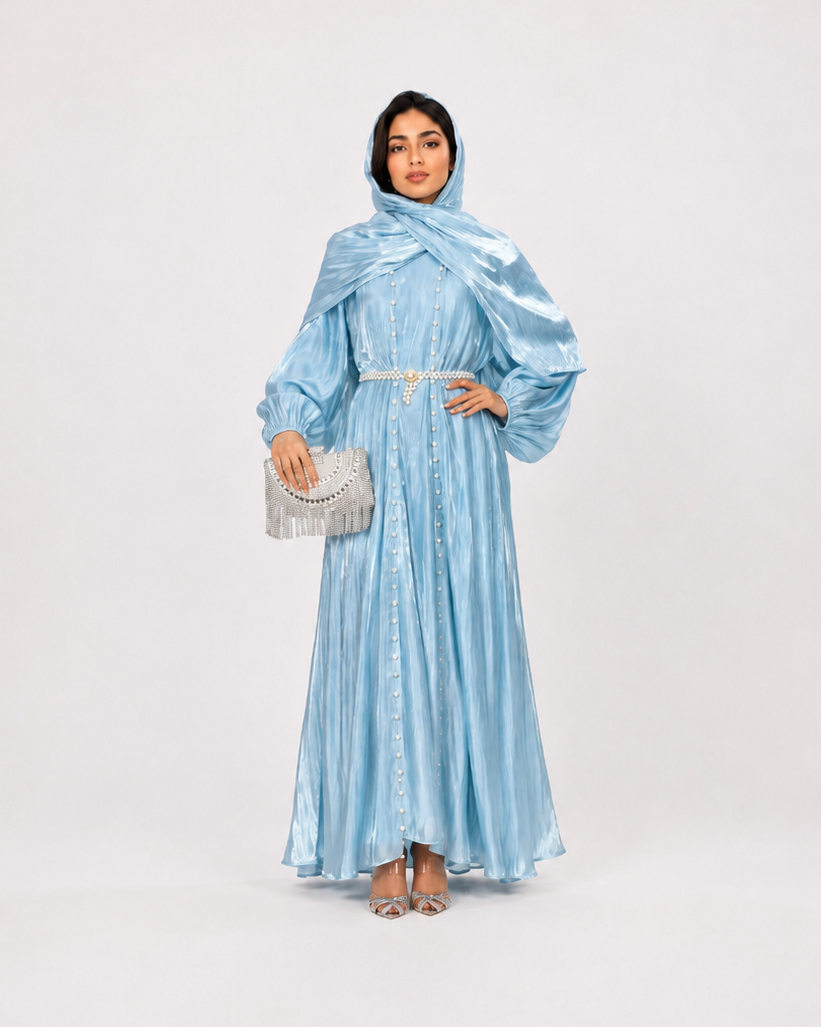 A woman modeling a light blue metallic maxi dress with a matching headscarf and holding a silver clutch, standing against a plain light gray studio background.
