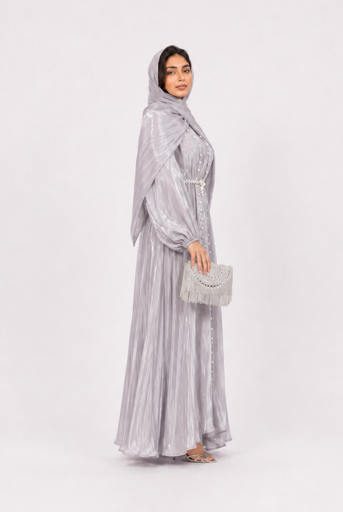 A woman modeling a light silver-gray maxi abaya with a matching headscarf and holding a small matching clutch bag, standing against a plain light gray studio background.