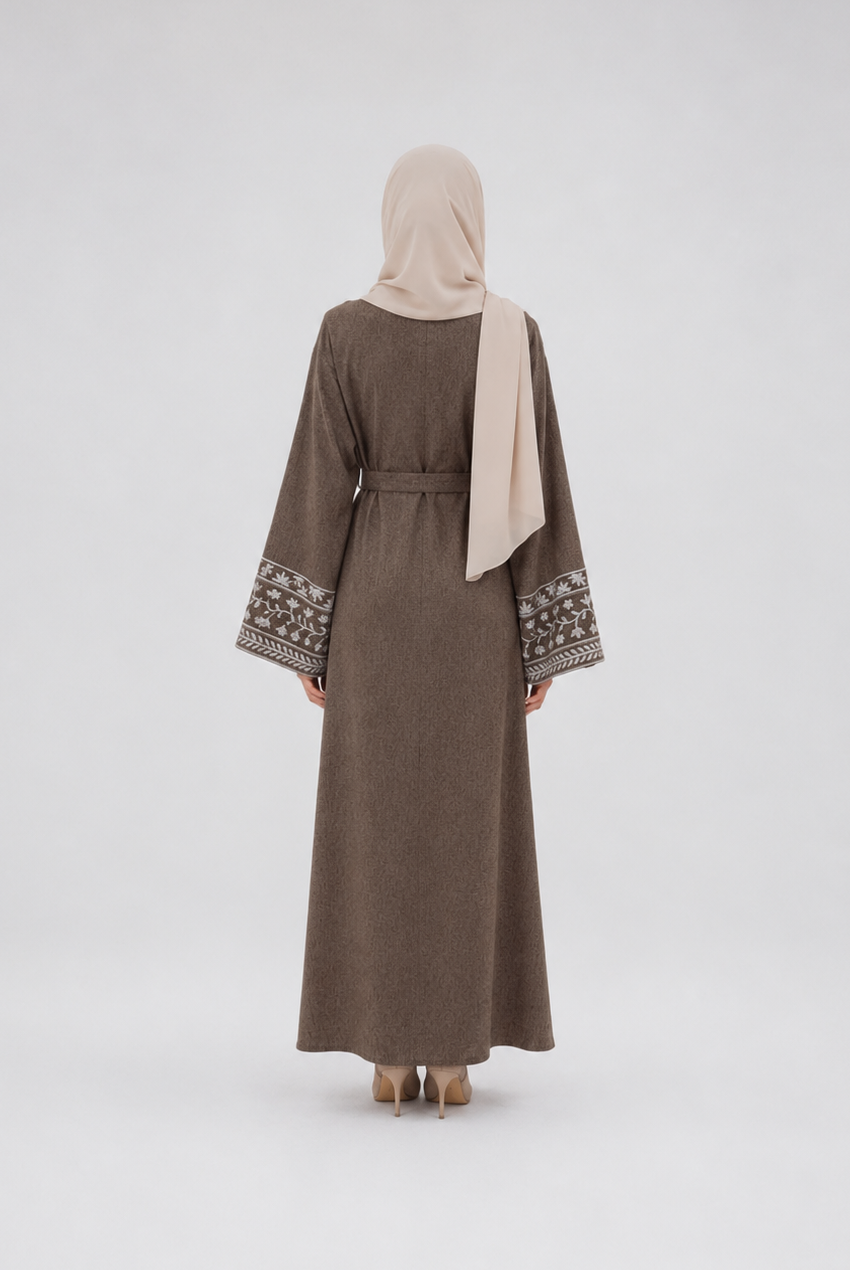 A woman viewed from behind wearing a long taupe-brown abaya with patterned embroidery on the wide sleeves and a light beige hijab, standing against a plain light gray studio background.