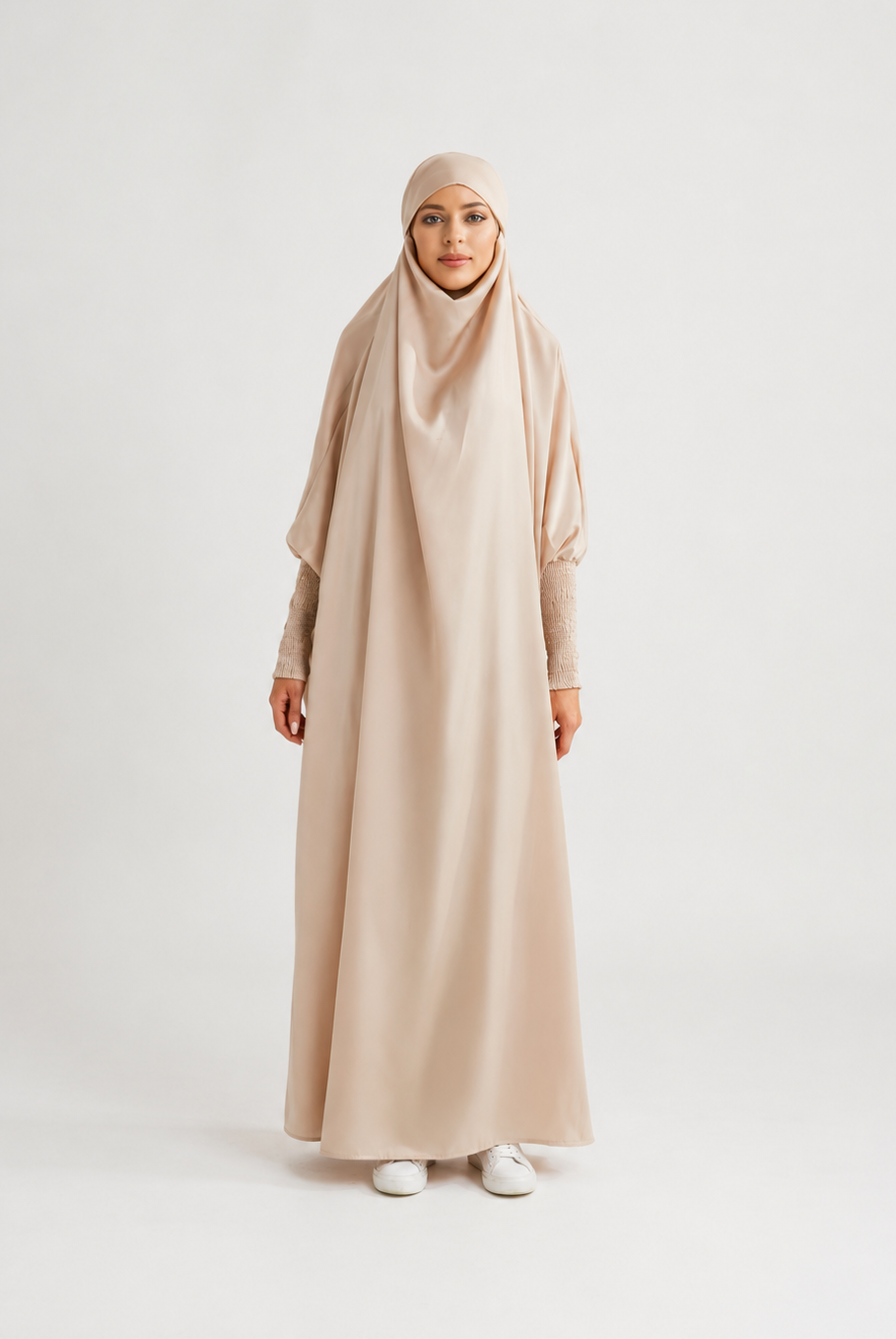 A woman wearing a long, flowing beige abaya dress with a matching hijab, standing against a plain light gray studio background.