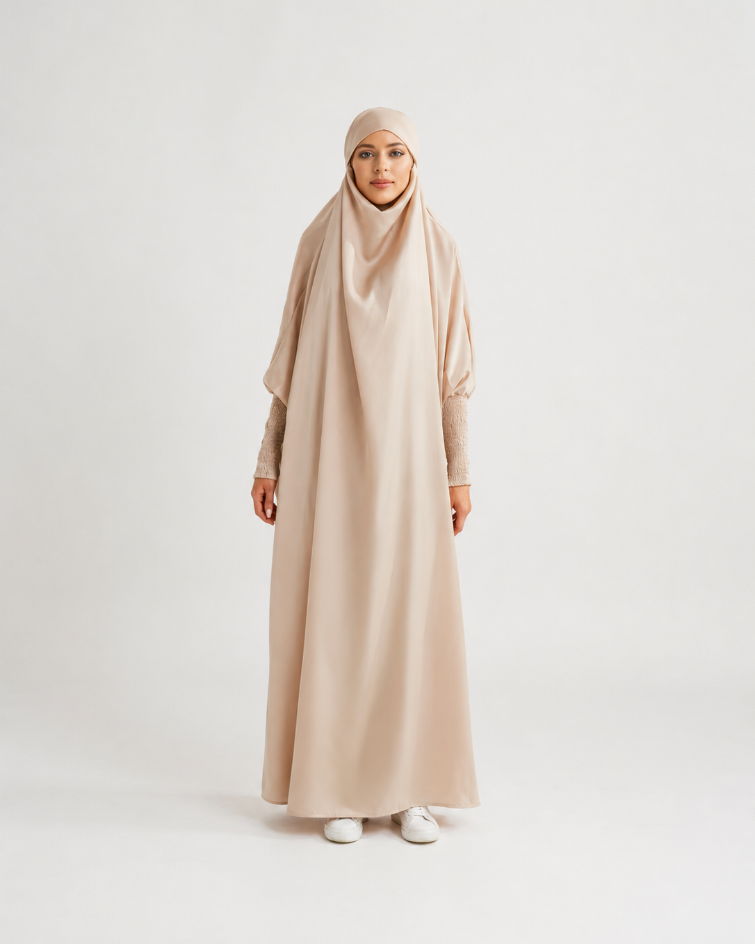 A woman wearing a long, flowing beige abaya dress with a matching hijab, standing against a plain light gray studio background.