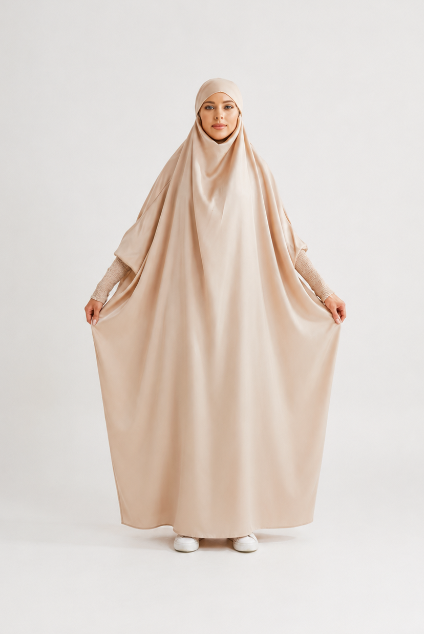 A woman wearing a loose, flowing beige hijab and long-sleeve abaya outfit, holding the fabric outwards against a plain light gray studio background.