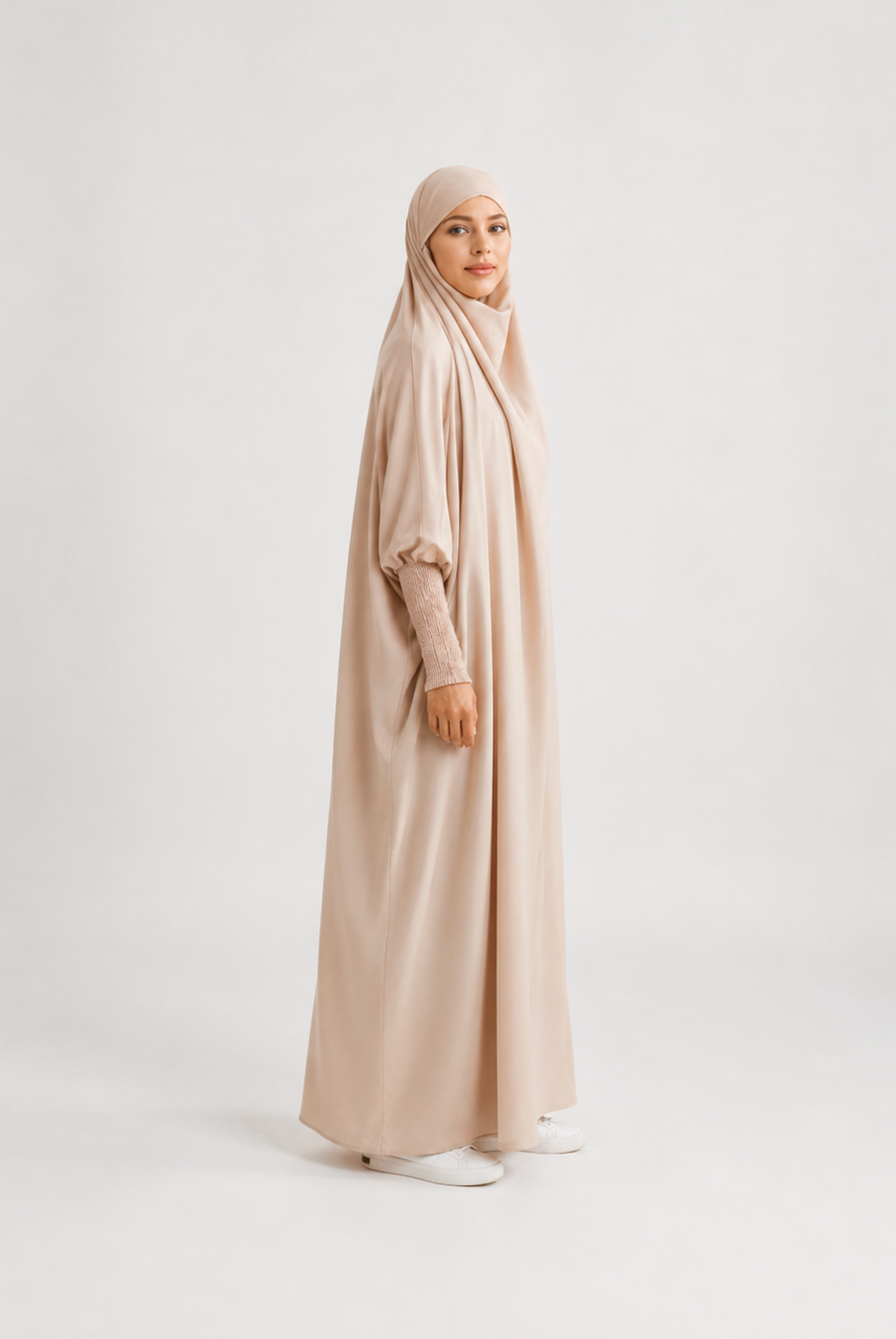 A woman wearing a long, flowing abaya in a light beige color with a matching hijab, standing in profile against a plain light gray studio background.