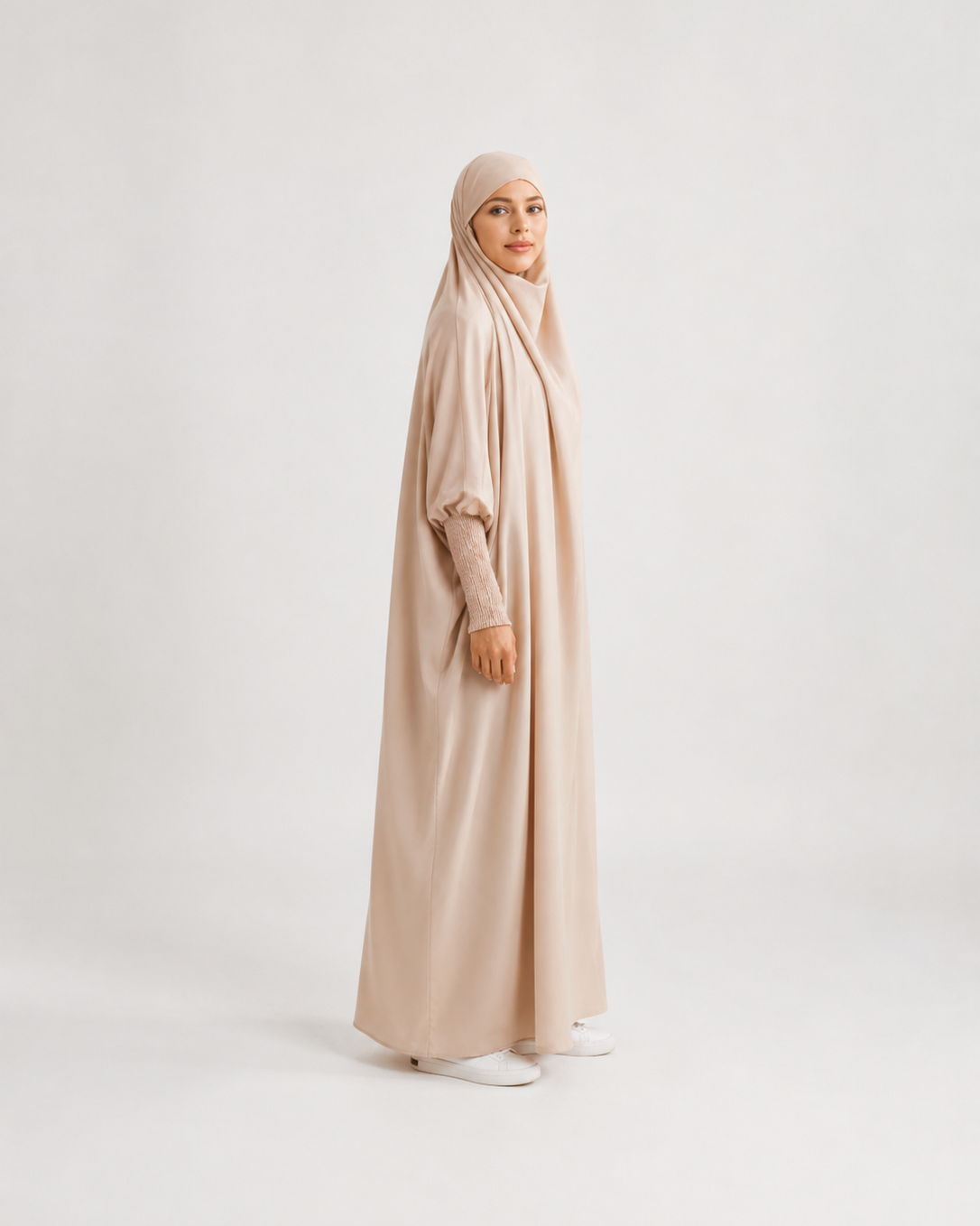 A woman wearing a long, flowing abaya in a light beige color with a matching hijab, standing in profile against a plain light gray studio background.