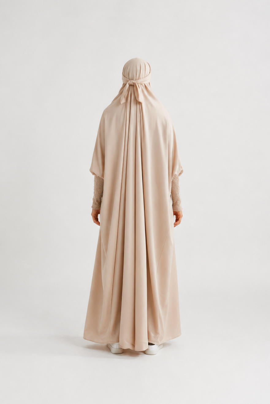 A person viewed from the back wearing a flowing light beige or blush-beige full-length garment with a matching head covering, standing against a plain light gray studio background.