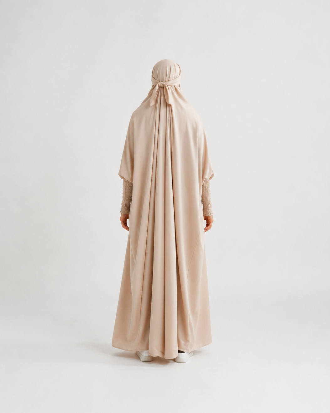 A person viewed from the back wearing a flowing light beige or blush-beige full-length garment with a matching head covering, standing against a plain light gray studio background.