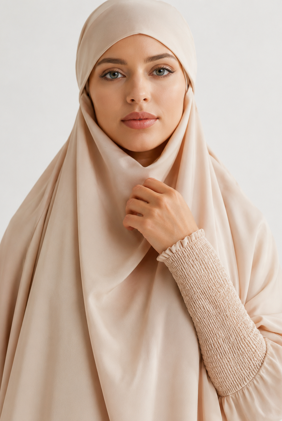 A woman wearing a light beige hijab and a matching long-sleeve top with a textured smocked sleeve, photographed against a clean light gray studio background.