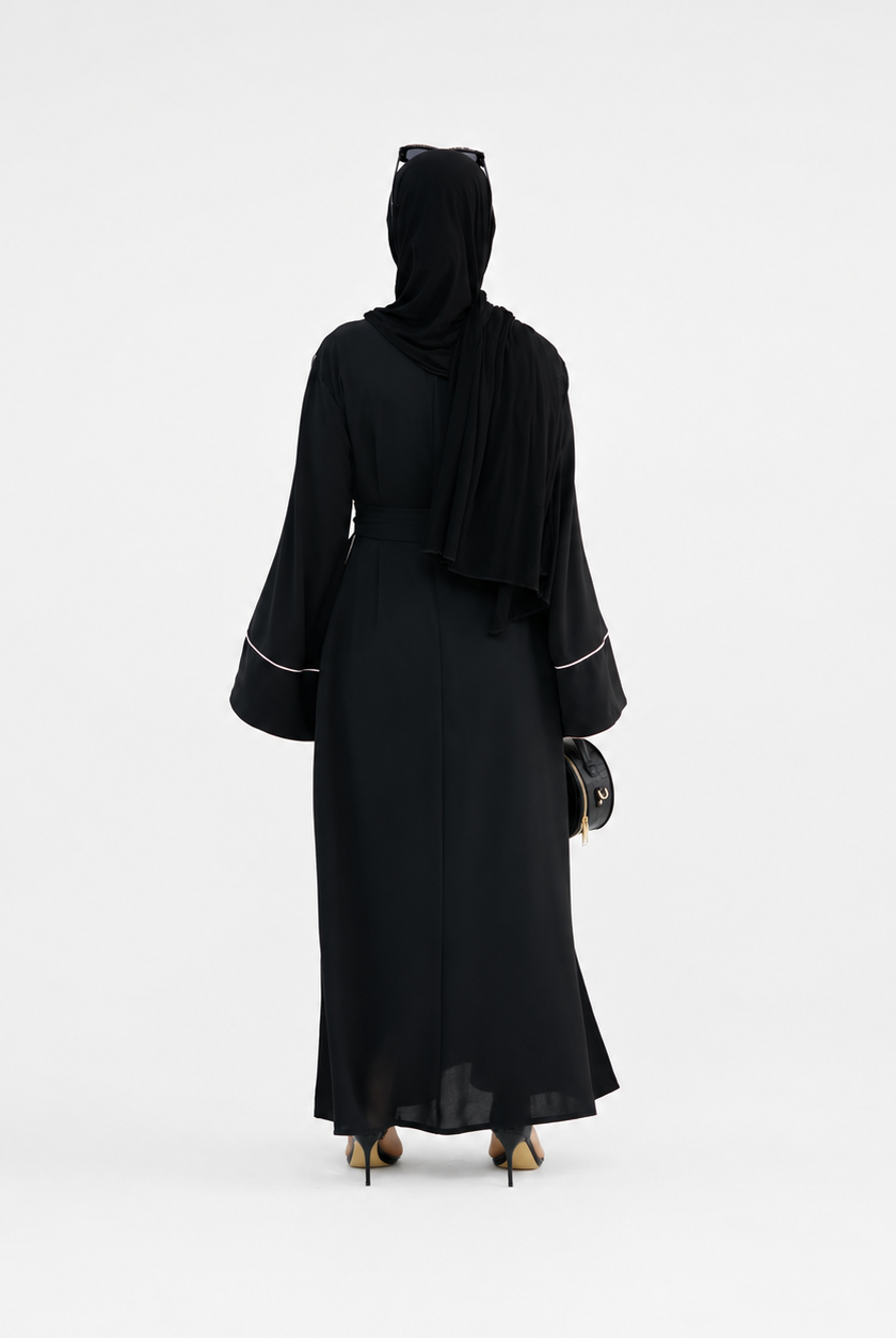 A woman viewed from behind wearing a long black abaya with wide sleeves and thin white piping details at the cuffs, along with a black hijab and black heels, against a plain white studio background.