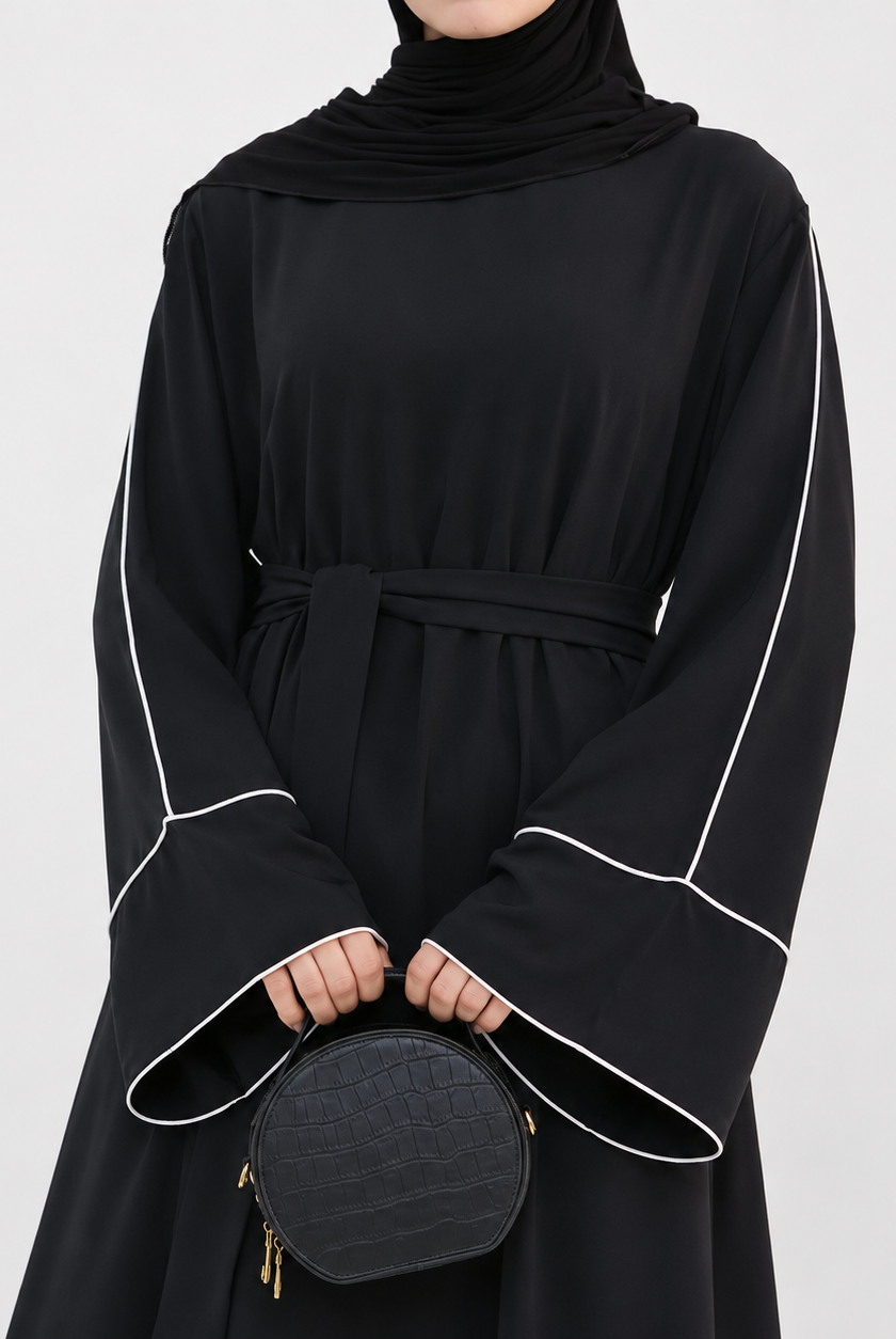 A person wearing a black long-sleeve abaya dress with white contrast piping and a matching waist tie, paired with a black headscarf and holding a small black quilted handbag against a light gray studio background.
