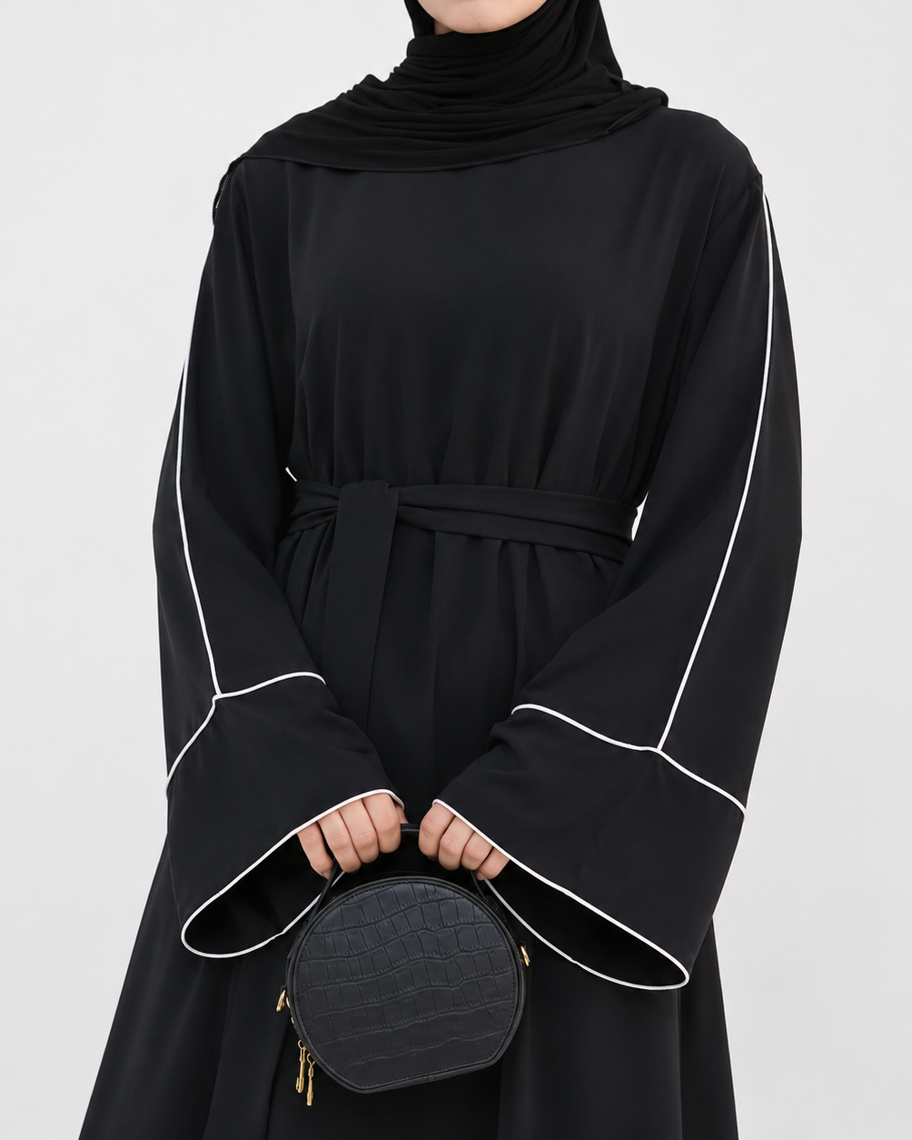 A person wearing a black long-sleeve abaya dress with white contrast piping and a matching waist tie, paired with a black headscarf and holding a small black quilted handbag against a light gray studio background.