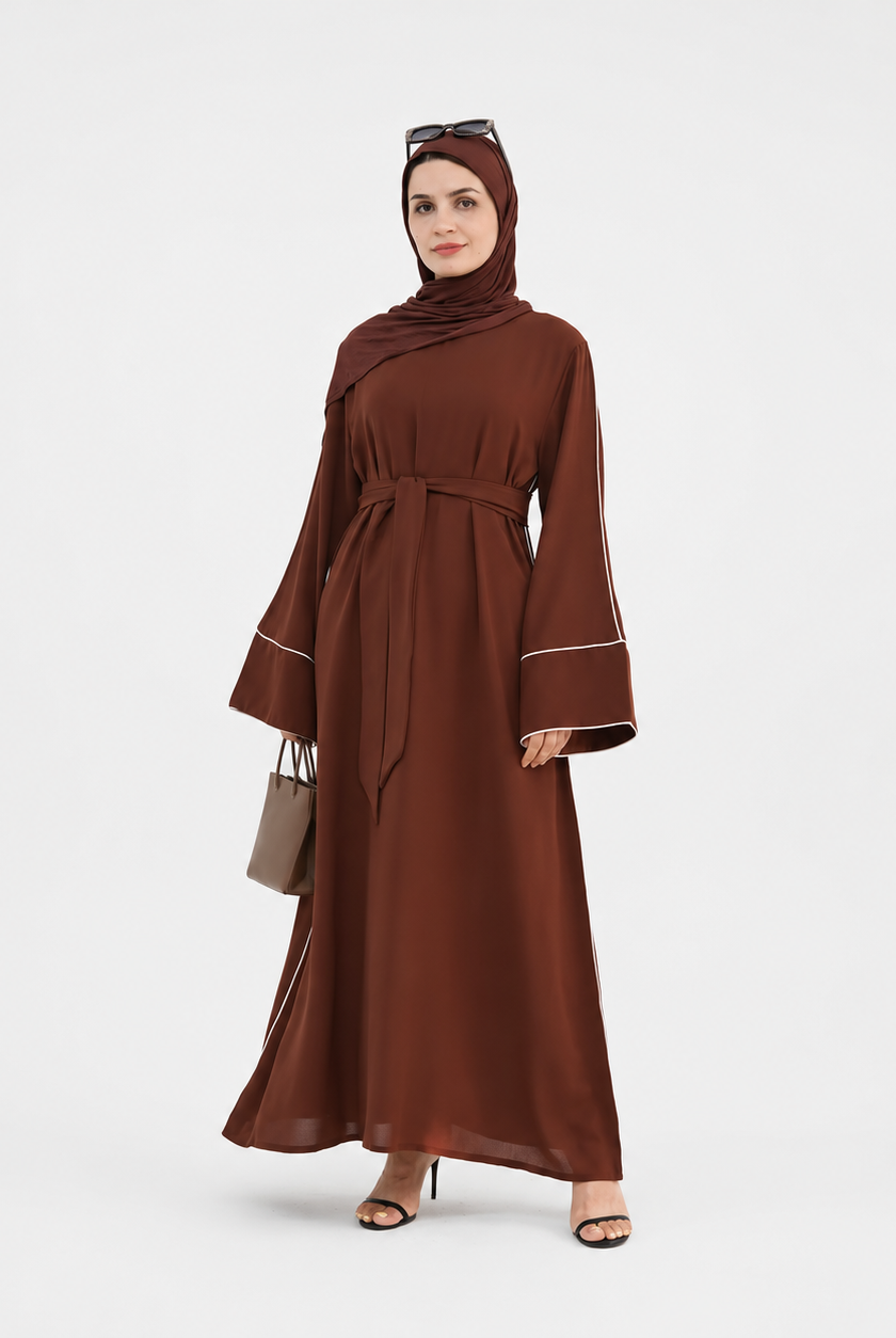 A woman modeling a long brown abaya dress with a matching headscarf, featuring wide bell sleeves with white piping details, a self-tie waist belt, and sunglasses on her head, standing against a light gray studio background.