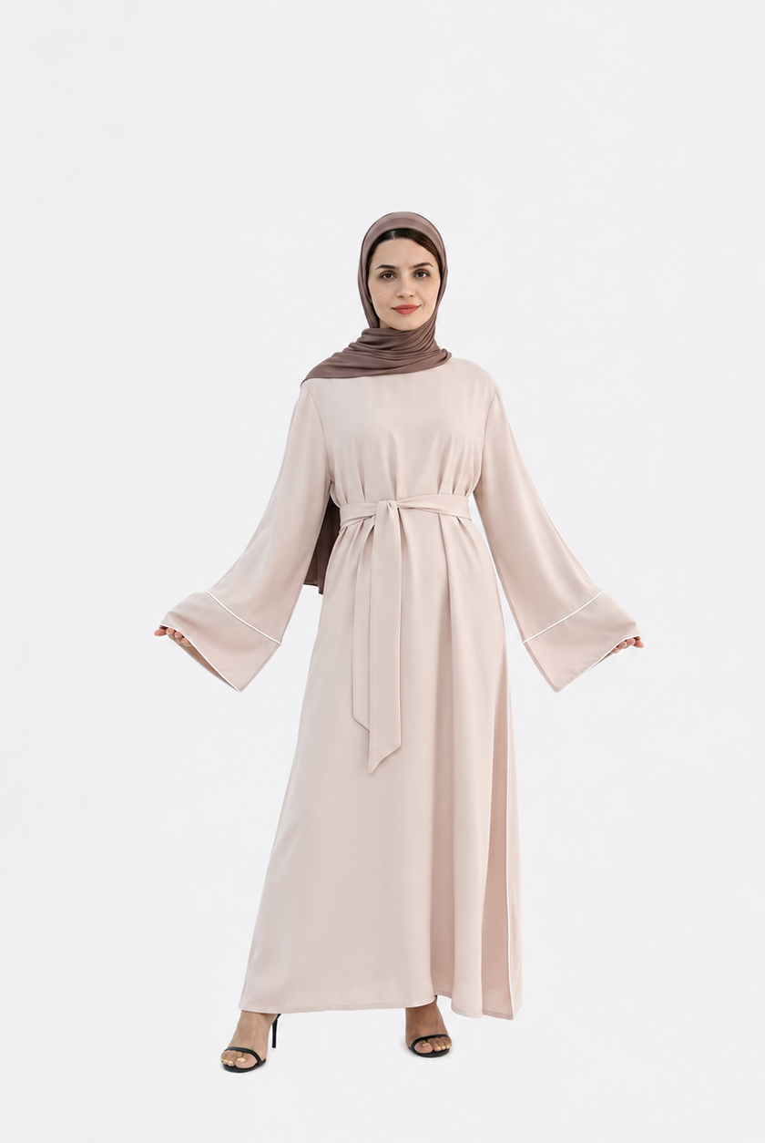 A woman modeling a long, light blush pink abaya dress with wide bell sleeves and a matching waist tie, paired with a brown hijab and black heeled sandals, set against a plain light gray background.