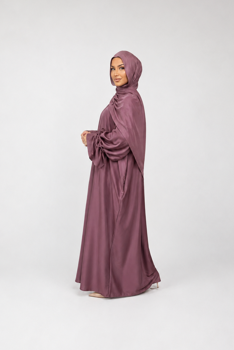 A woman wearing a flowing mauve-colored abaya and matching hijab, standing in profile against a light gray studio background.
