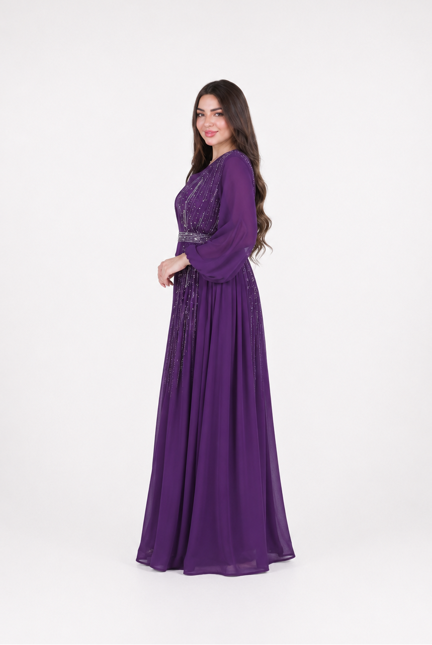 A woman modeling a long, floor-length purple gown with long sleeves and a fitted waist, standing against a plain light gray studio background.