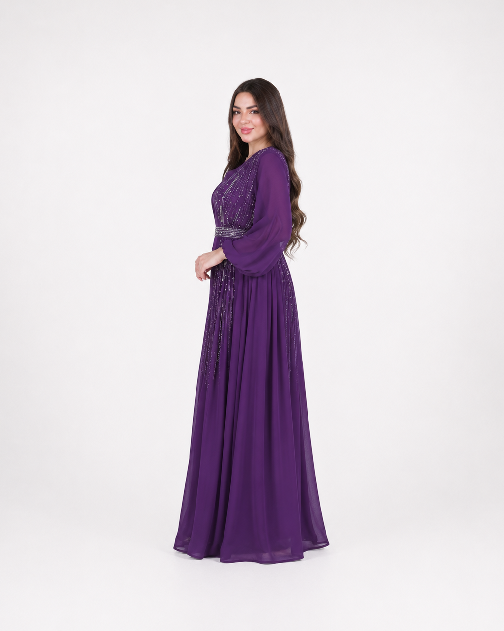 A woman modeling a long, floor-length purple gown with long sleeves and a fitted waist, standing against a plain light gray studio background.