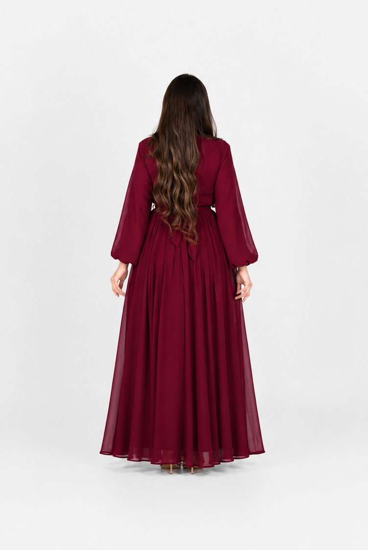 A woman viewed from behind wearing a long, flowing burgundy dress with three-quarter sleeves, standing against a plain light gray studio background.