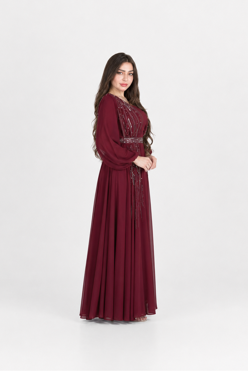 A woman modeling a long, floor-length maroon dress with long sleeves, a fitted waist, and embroidered detailing on the bodice, standing against a plain light gray studio background.