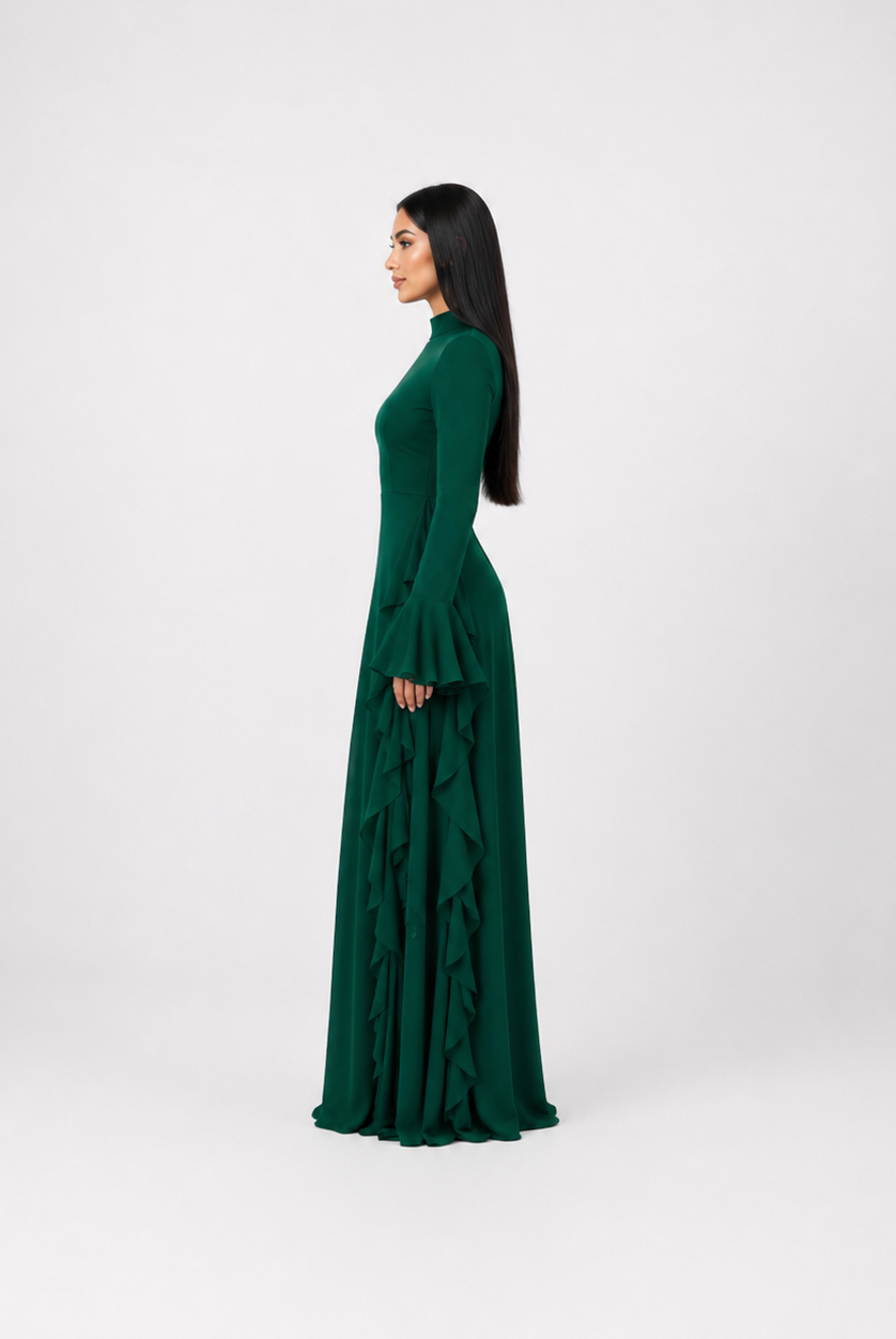A woman in profile wearing a long-sleeved, high-neck, floor-length emerald green dress with a ruffled skirt, standing against a light gray studio background.