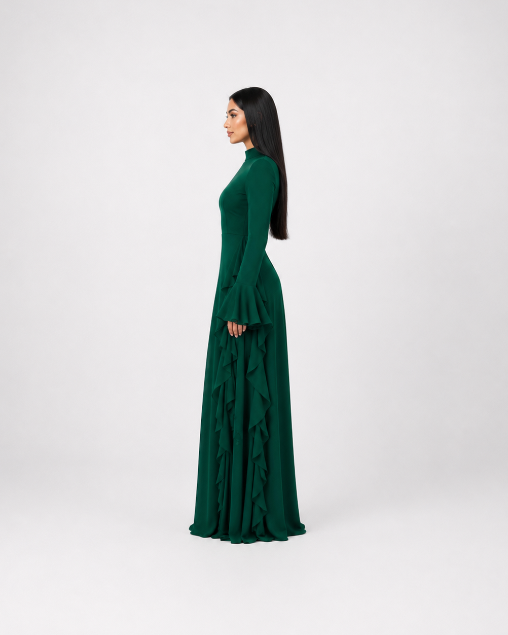 A woman in profile wearing a long-sleeved, high-neck, floor-length emerald green dress with a ruffled skirt, standing against a light gray studio background.