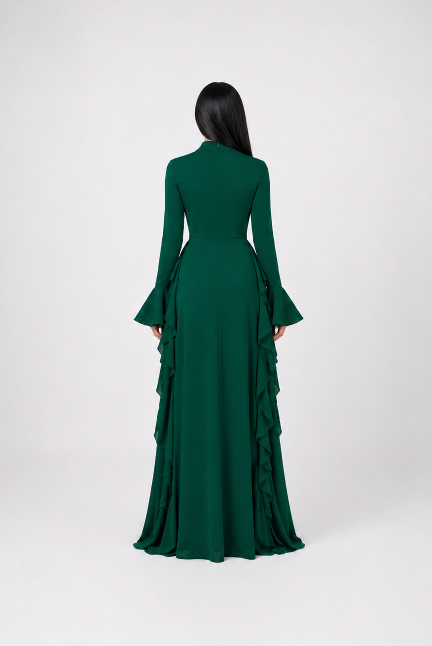A woman viewed from the back wearing a long-sleeved emerald green gown with a structured waist and layered ruffle detailing along the skirt, set against a plain light gray studio background.