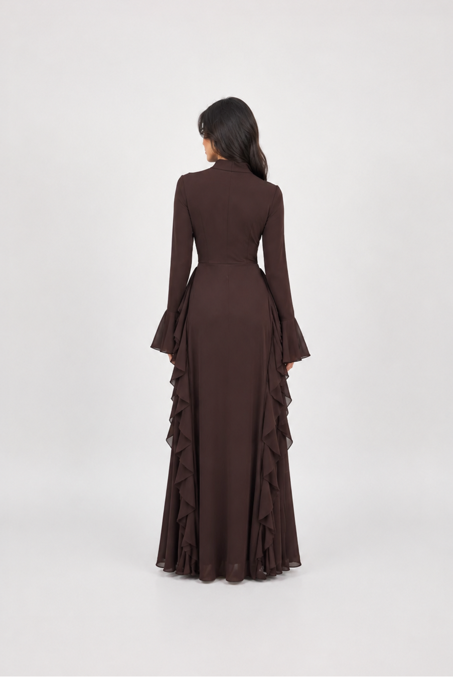 A woman viewed from behind wearing a long-sleeved dark brown maxi dress with flared cuffs and layered ruffle detailing along the skirt, set against a plain light gray studio background.