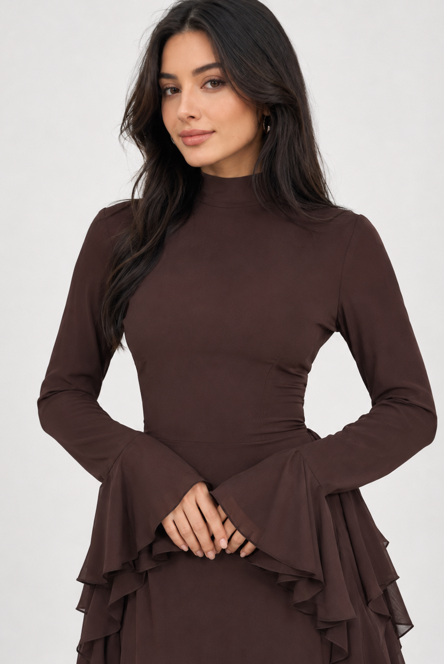 A woman wearing a dark brown long-sleeve mock neck dress with flared cuffs and a tiered ruffle hem, photographed against a light gray studio background.