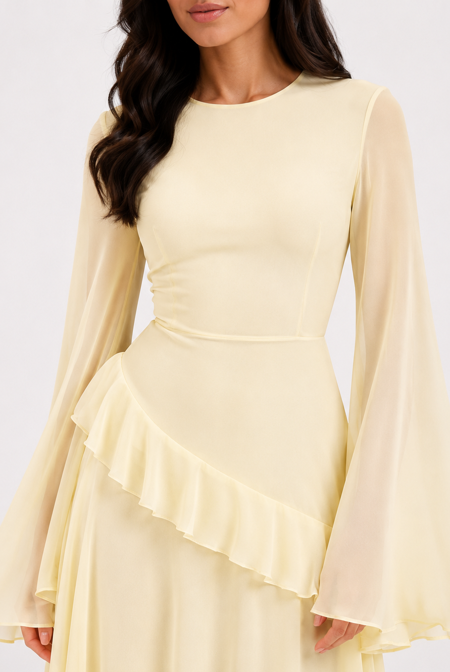 A woman wearing a pale yellow long-sleeve dress with a fitted bodice, ruffled waist detail, and flowing sheer bell sleeves against a plain light background.