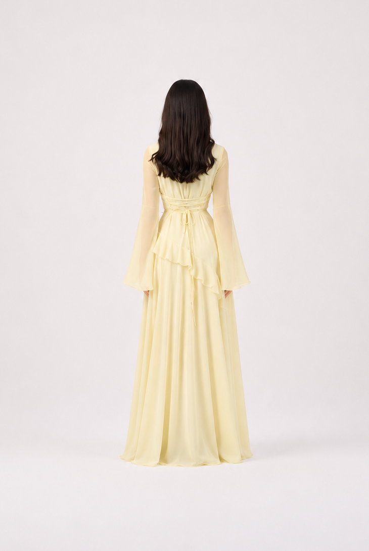 A woman viewed from behind wearing a pale yellow long-sleeve maxi dress with a ruffled waist, standing against a plain light gray studio background.