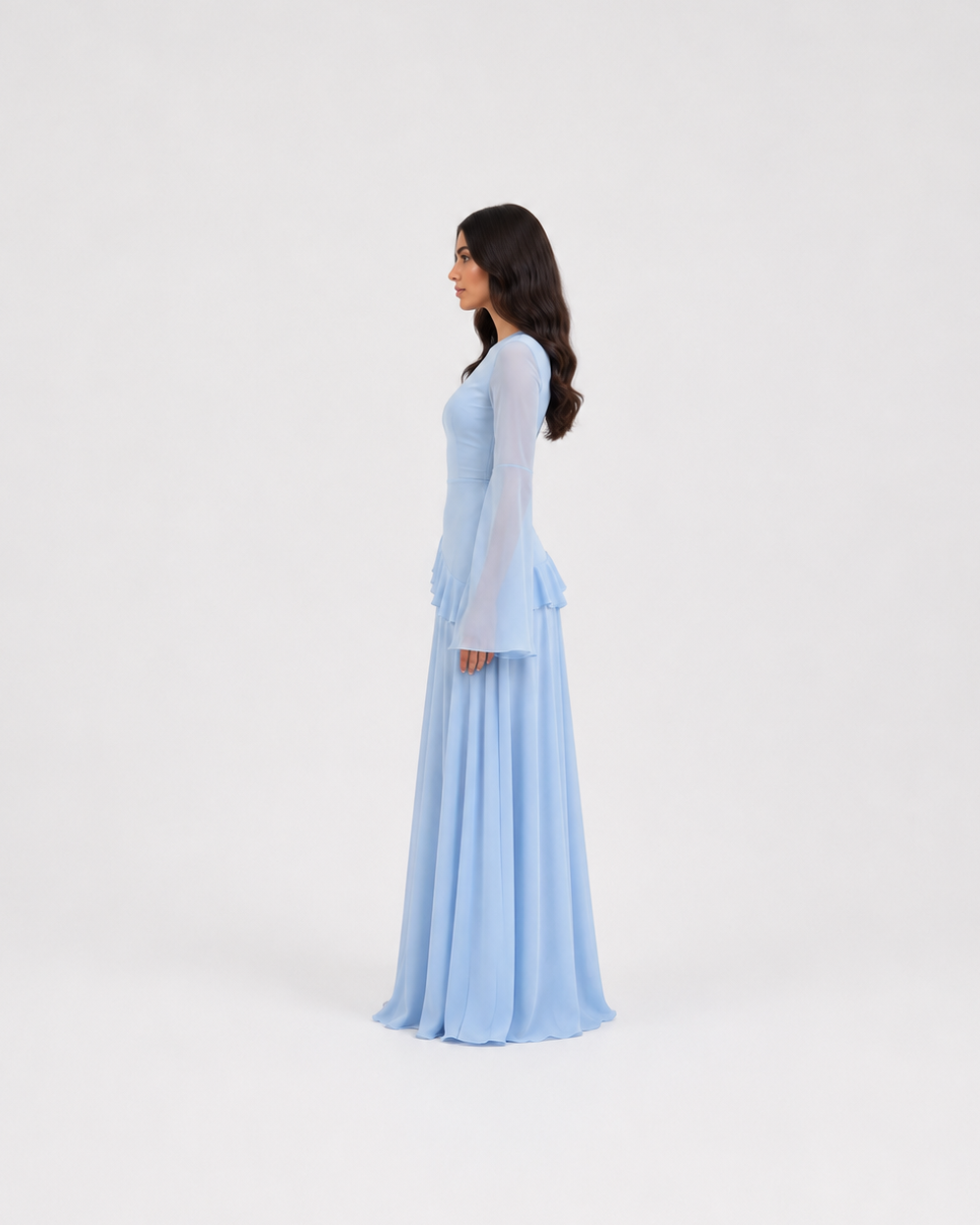A woman wearing a light blue long-sleeve maxi dress with a fitted bodice and flowing skirt, shown in profile against a plain light gray studio background.