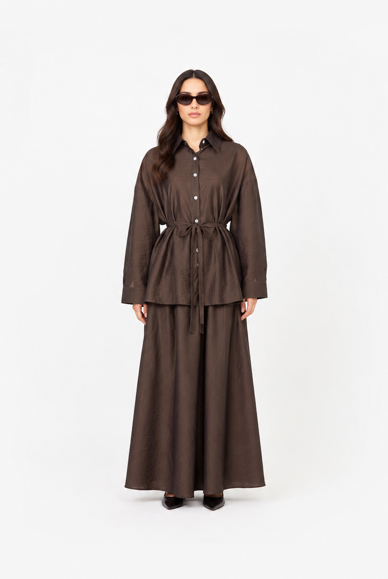 A model wearing a dark brown two-piece outfit consisting of a long-sleeve button-front shirt jacket with a tie waist and a matching long skirt, standing against a plain white studio background.