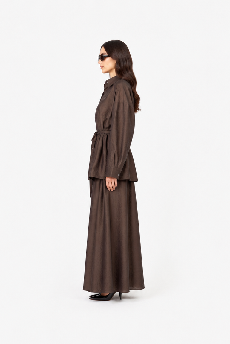 A woman in side profile wearing a dark brown belted long-sleeve jacket and matching wide-leg skirt against a plain white studio background.