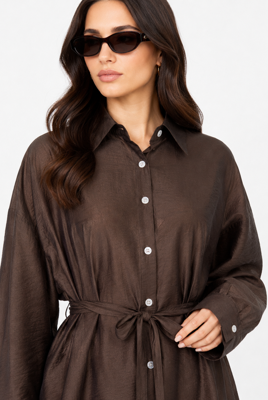 A woman wearing a dark brown long-sleeve button-up shirt dress with a tie waist belt, styled with black sunglasses against a plain white background.
