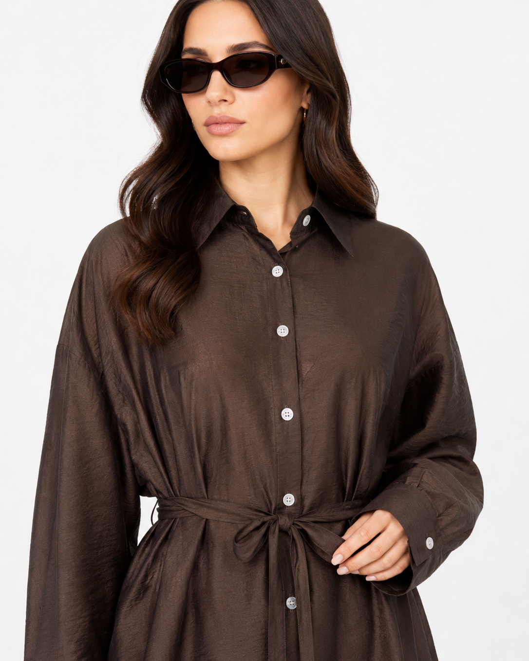 A woman wearing a dark brown long-sleeve button-up shirt dress with a tie waist belt, styled with black sunglasses against a plain white background.