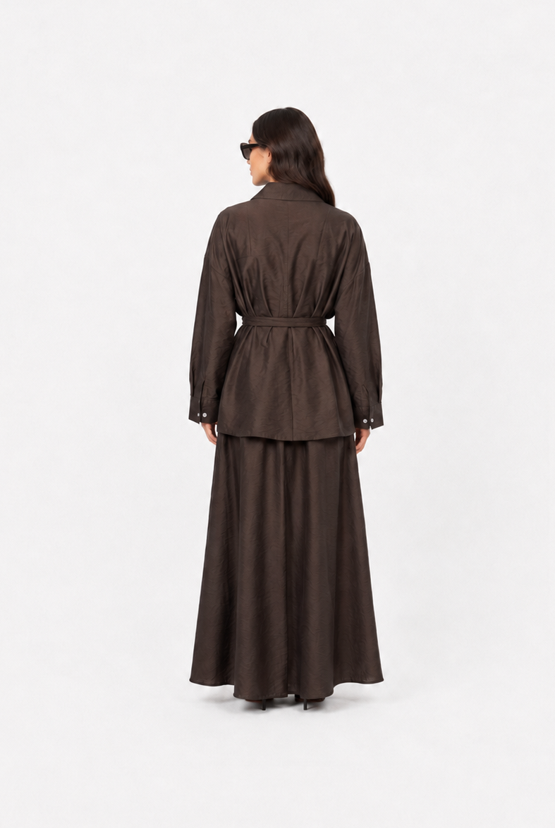 A model viewed from behind wearing a dark brown long-sleeved jacket with a matching waist tie and a long, flowing skirt against a plain white studio background.