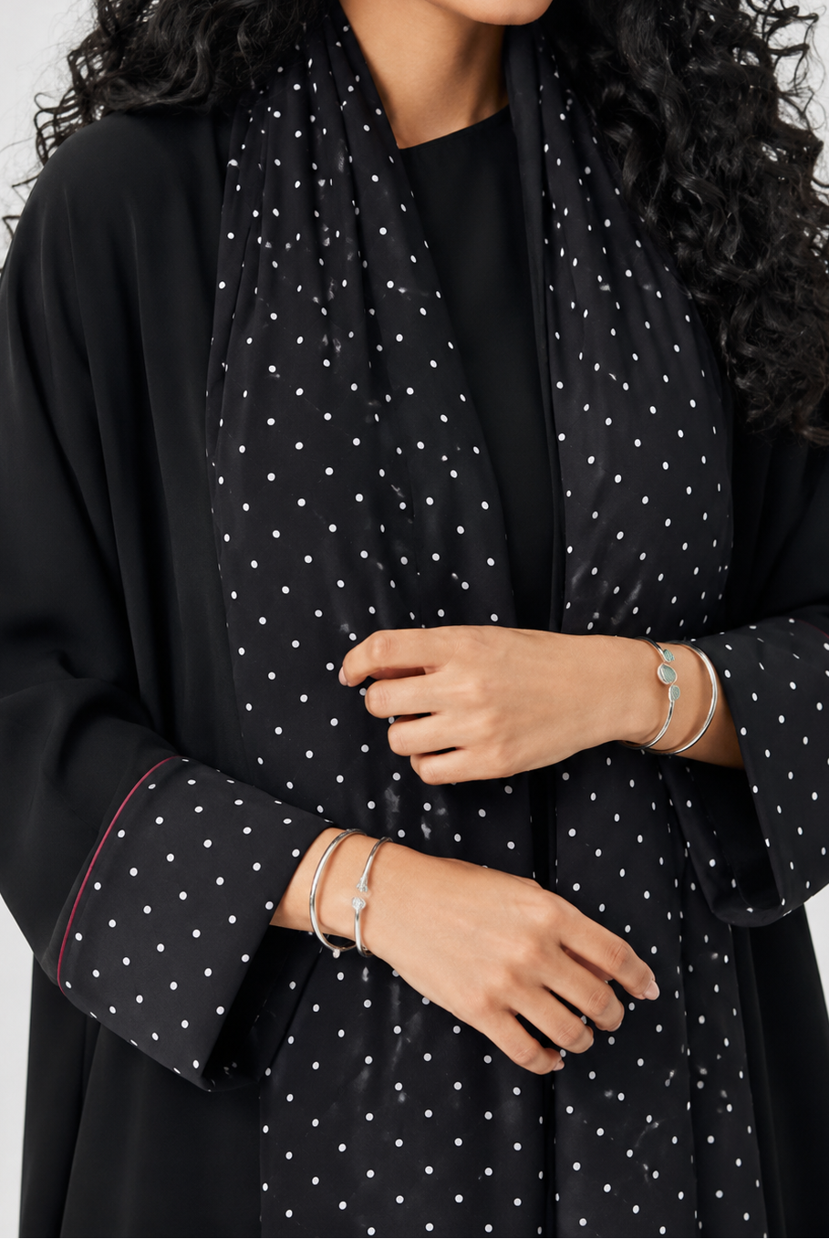 A close-up of a woman wearing a black long-sleeve abaya or robe with a white polka-dot patterned scarf, shown against a plain light gray background.
